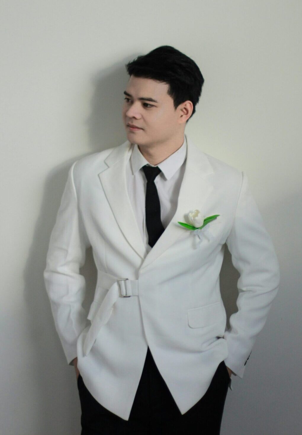 Elegant groom in white suit with black tie and boutonniere looking to the side indoors.